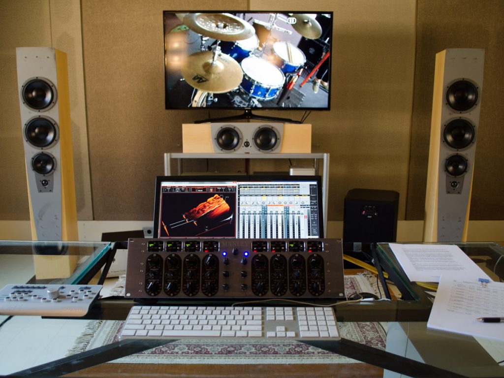 Vista frontal. TV entre duas caixas acusticas. Imagem de bateria na tela. Mesa com teclado, equalizador, computador.